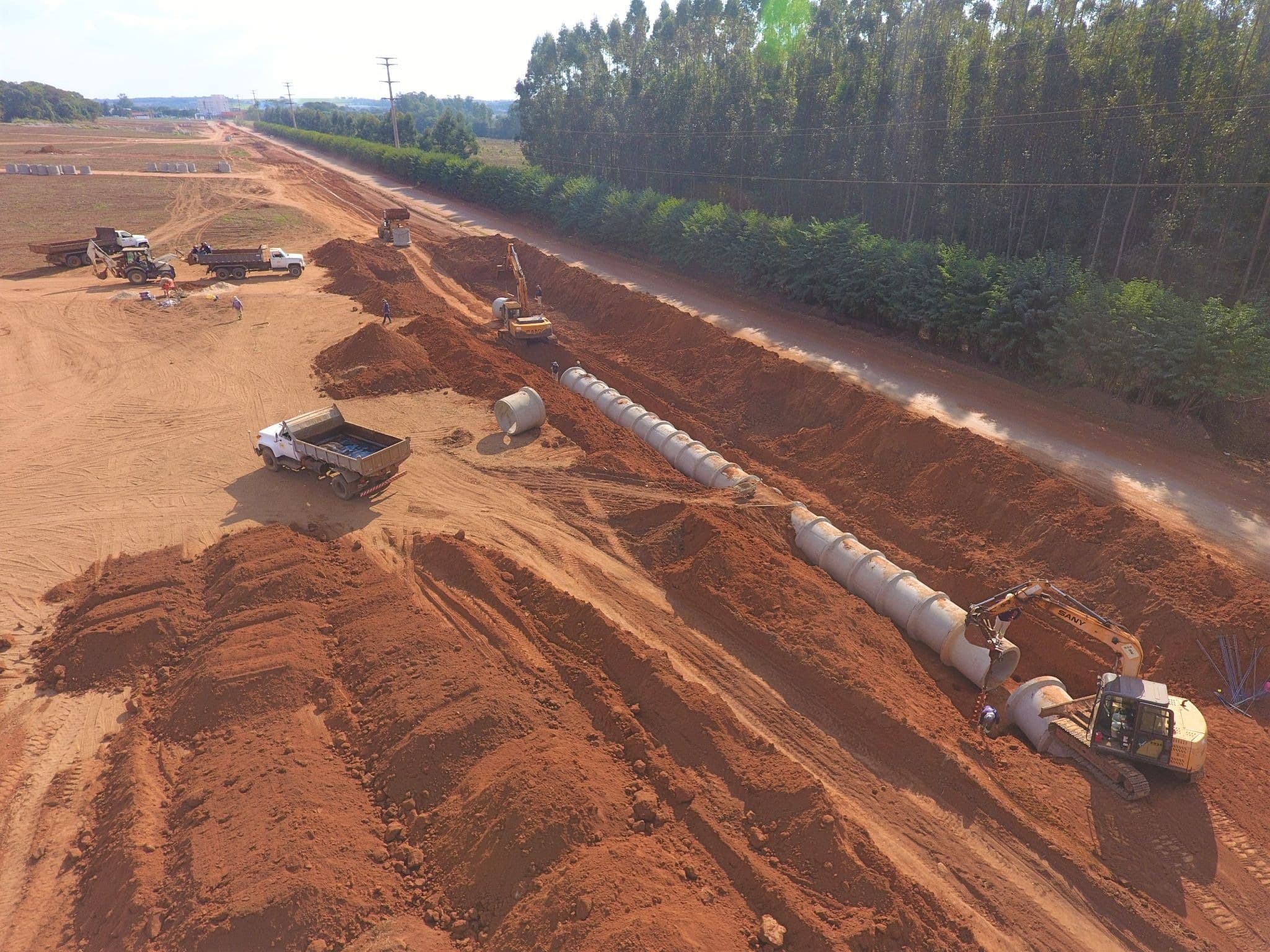 Vista de uma obra de terraplenagem com escavadeiras e instalação de tubulação de concreto.